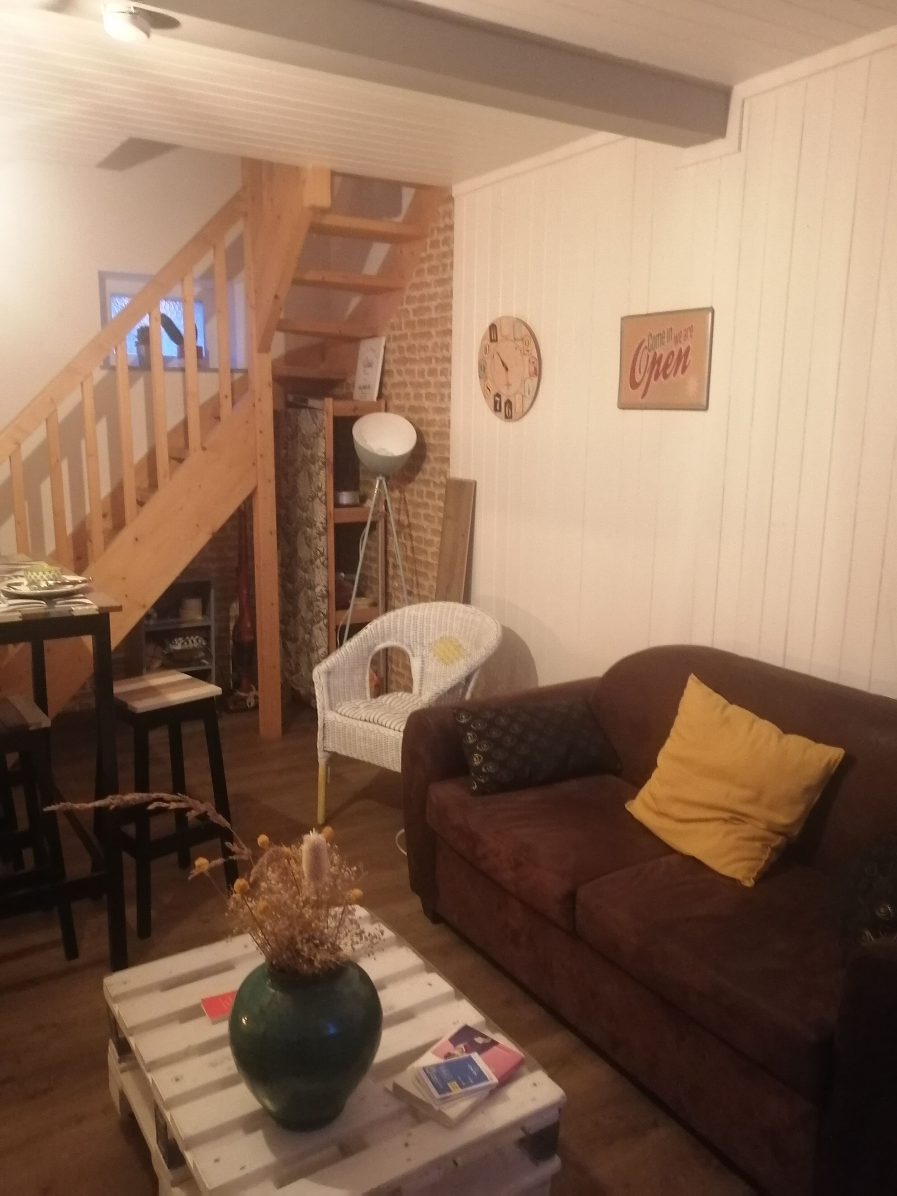 Un salon avec un canapé marron, un fauteuil blanc, un escalier en bois et une horloge au mur.