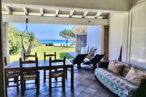 Überdachte Terrasse mit Tisch und Stühlen, einer Couch und einem Hängesessel mit Blick auf einen Strand.