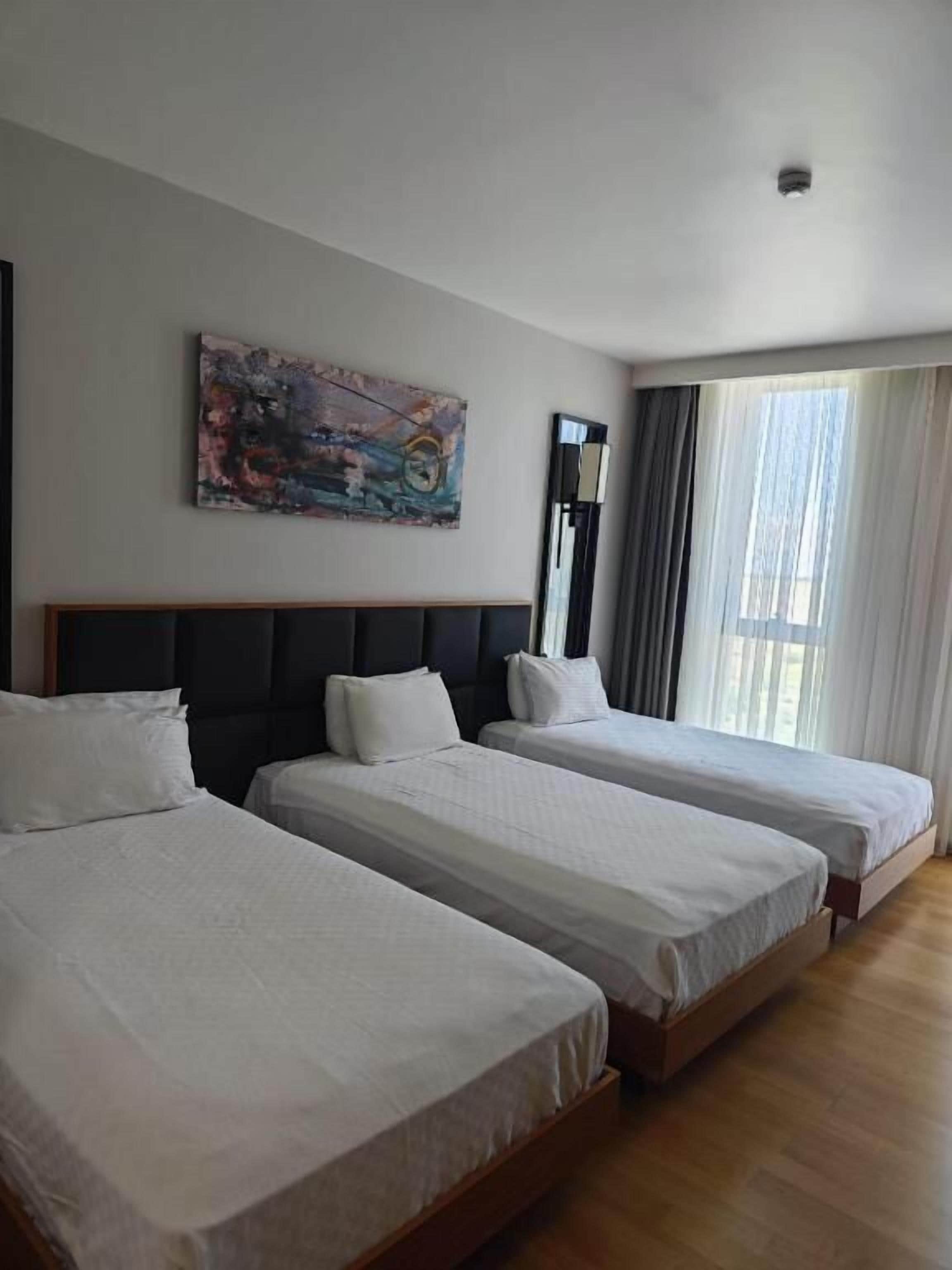 A hotel room with three single beds, a large window with curtains, and a painting on the wall.