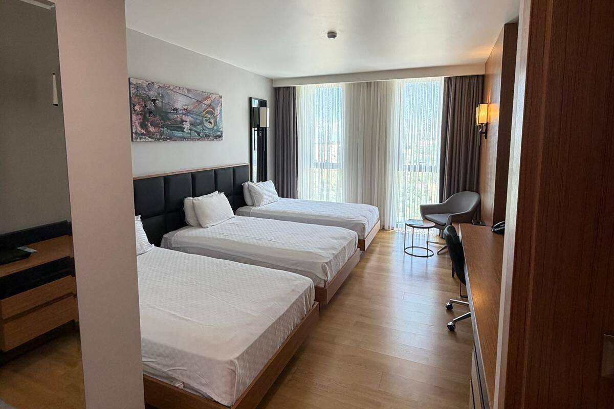Ein Hotelzimmer mit drei Betten, einem Schreibtisch, einem Stuhl und einem großen Fenster mit Vorhängen.