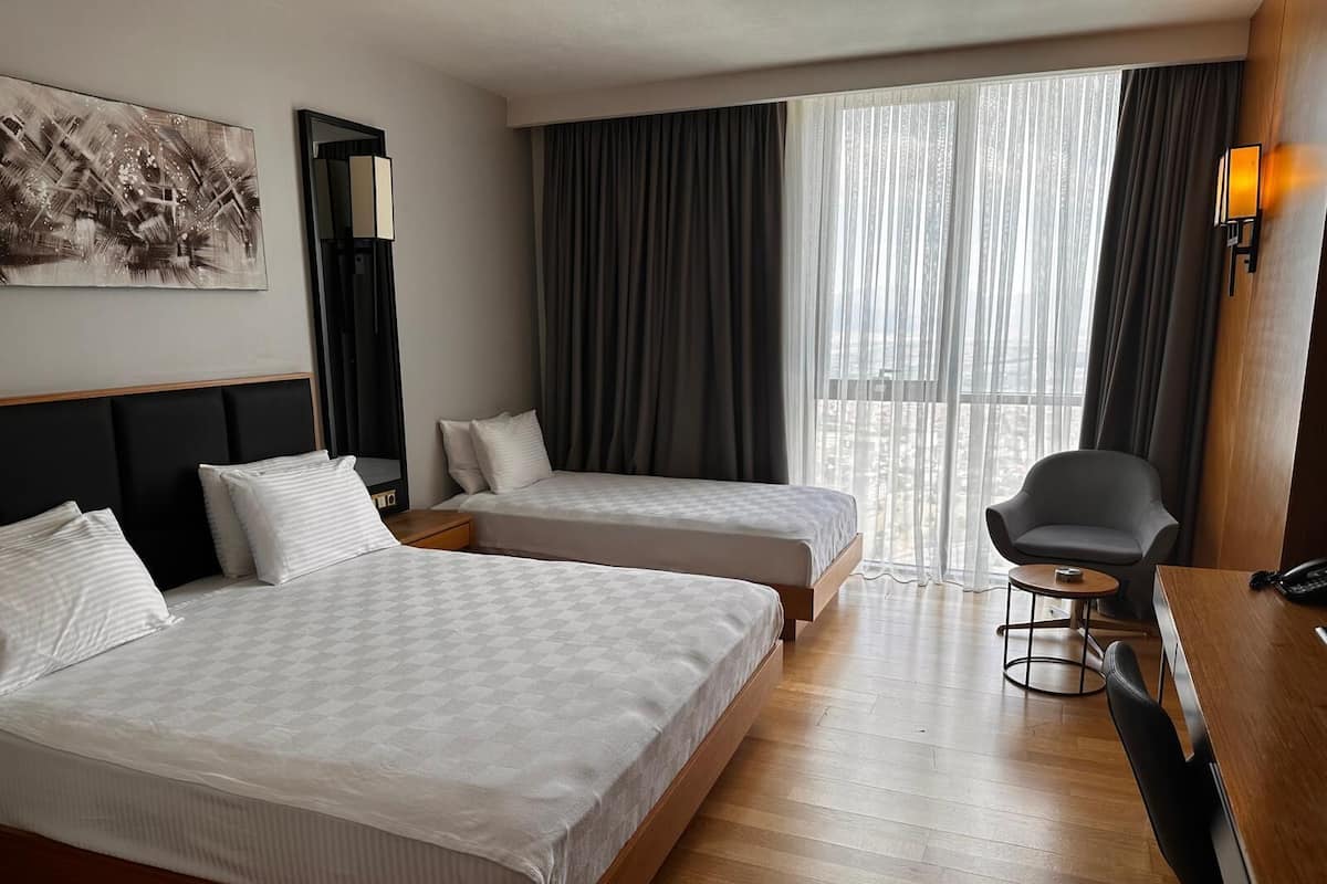 Ein Hotelzimmer mit zwei Betten, einem Schreibtisch, einem Stuhl, einer Lampe und einem großen Fenster mit Vorhängen.