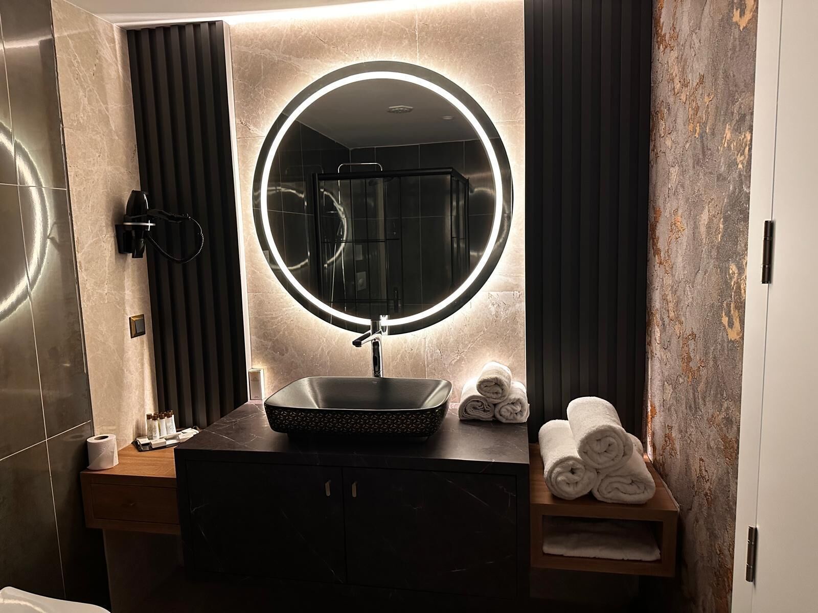 A modern bathroom with a round mirror, dark vanity, and towels on a shelf.