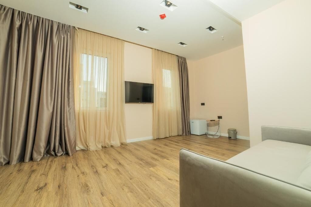 A room with a bed, a TV, a window with curtains, a ceiling light, and a small table with a trash bin.