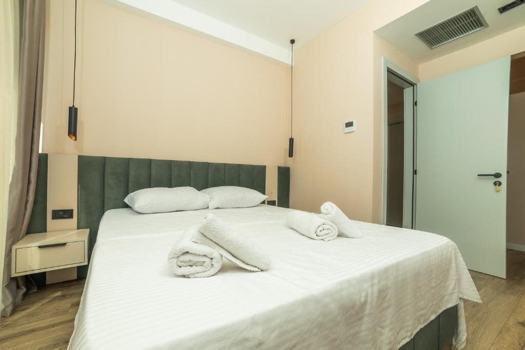 A neatly made bed with white linens and rolled towels, a bedside table, a green upholstered headboard, and a light-colored wall.