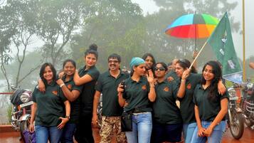 A group of people in matching shirts posing with a colorful umbrella and a flag, with trees and a motorcycle in the background.