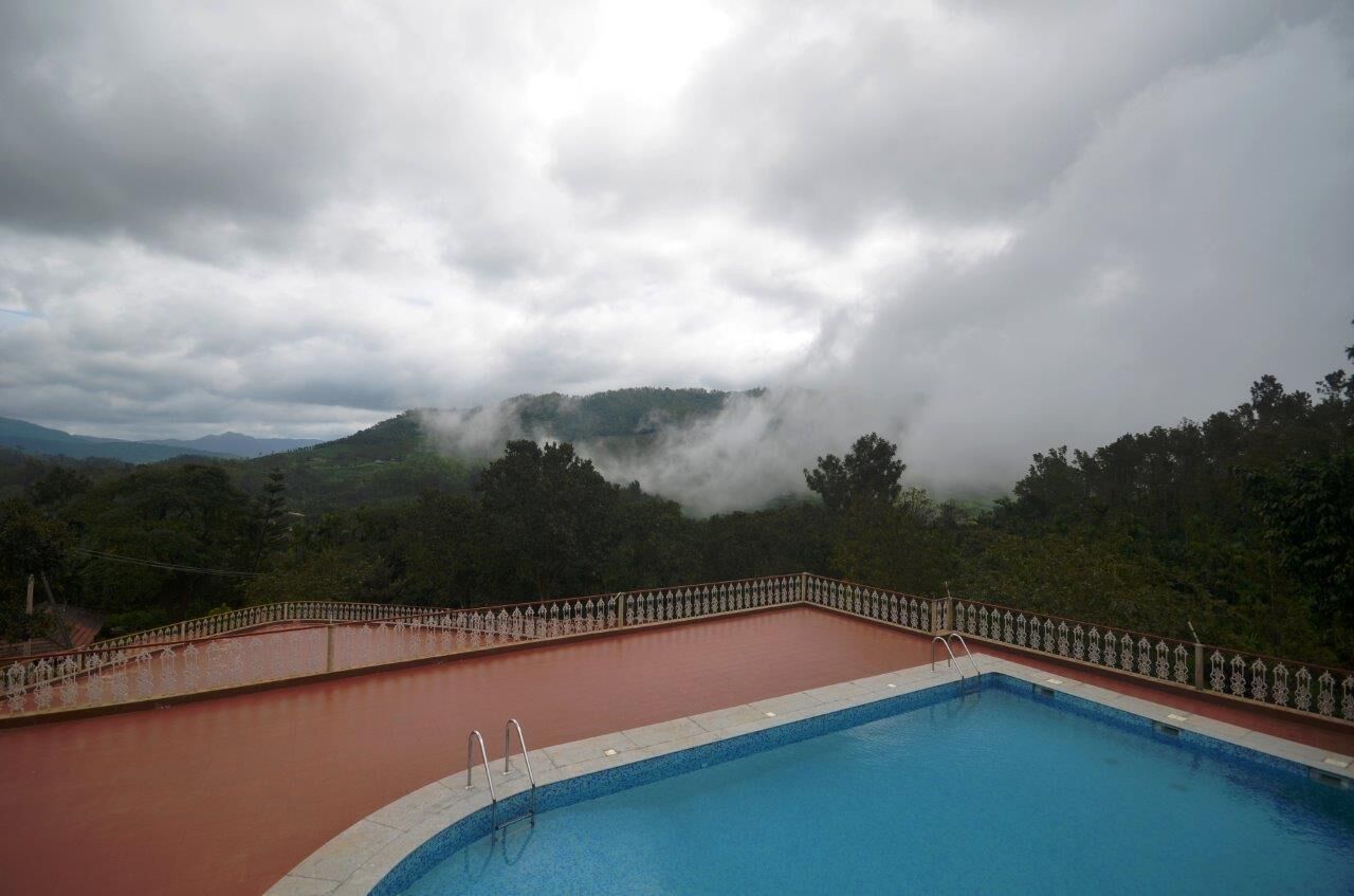 A swimming pool with a red and blue color scheme, surrounded by a decorative fence, set against a backdrop of mist-covered hills and a cloudy sky.