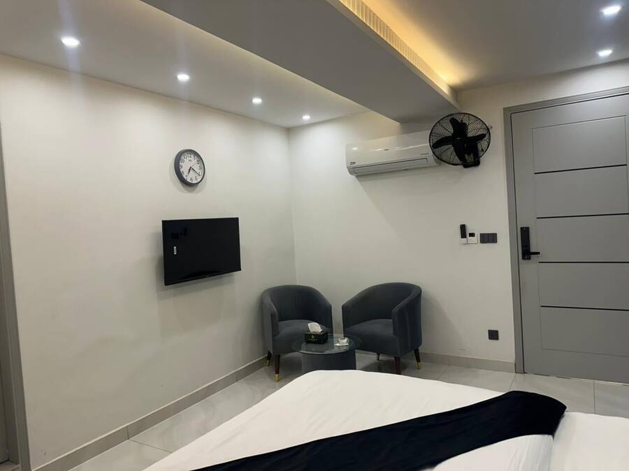 Et moderne hotelværelse med en seng, to lænestole, et lille bord, et tv ophængt på væggen, en aircondition-enhed og en dør.