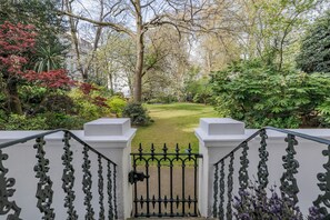 Une entrée fermée par un portail mène à un jardin avec une pelouse, des arbres et un bâtiment blanc en arrière-plan.