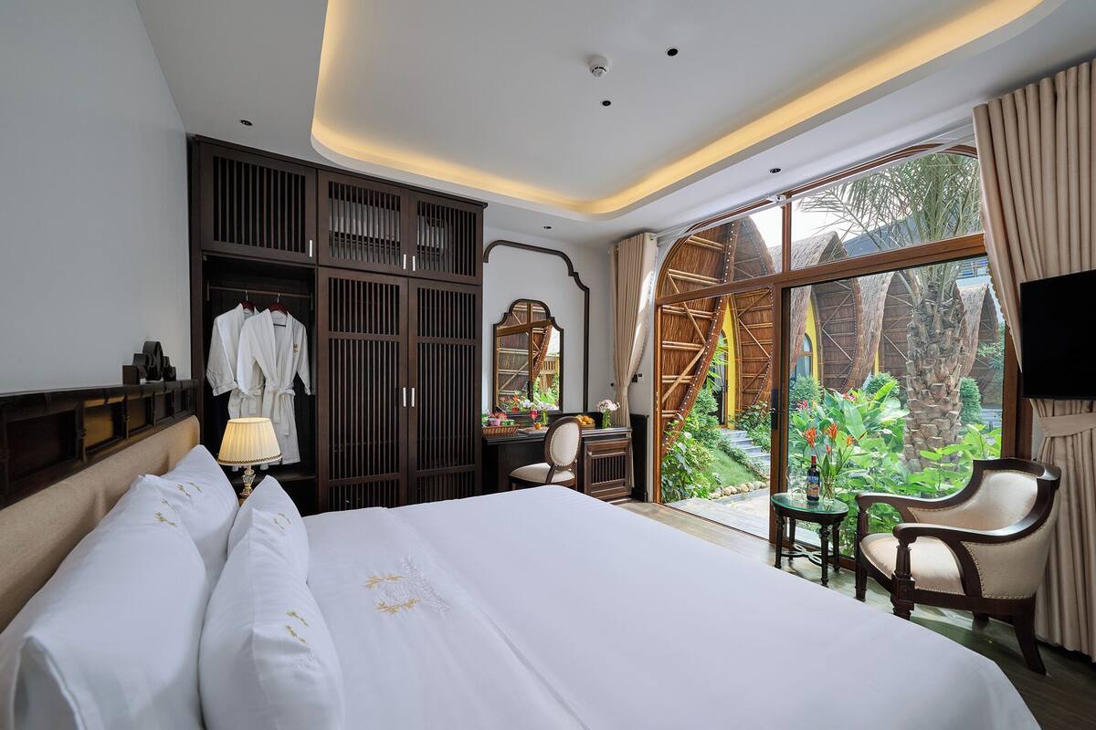 A hotel room with a large bed, a desk, a chair, a wardrobe, and a view of a garden.