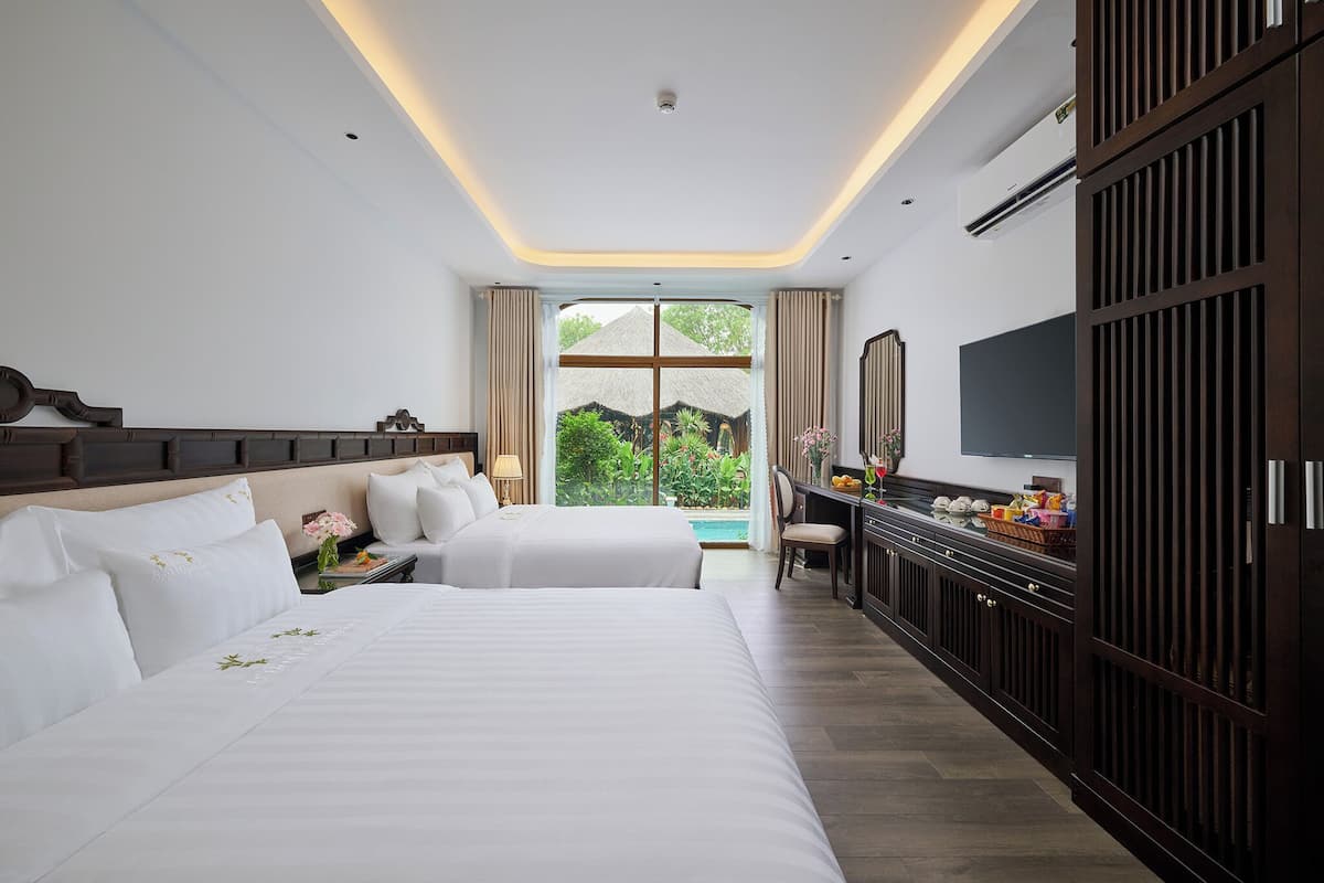 A hotel room with two beds, a TV, a desk, and a view of greenery.