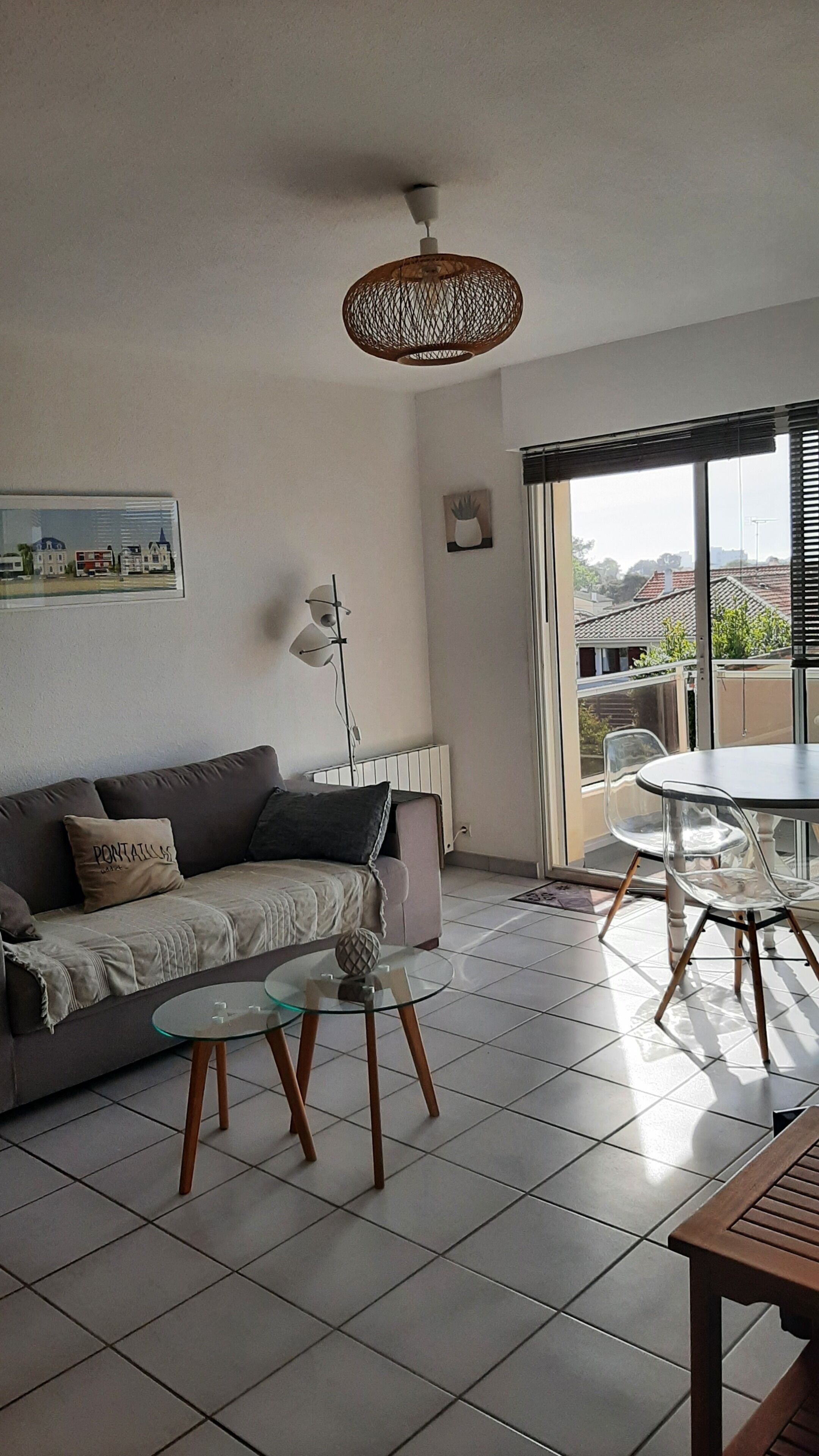 A living room with a sofa, a glass coffee table, a dining table with chairs, a hanging light fixture, and a window with blinds.