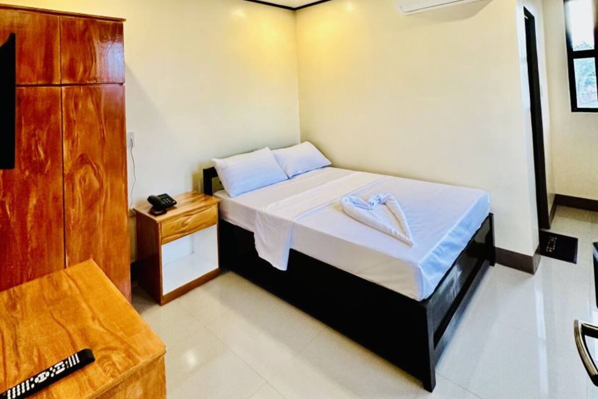 A hotel room with a bed, bedside table, wooden wardrobe, and an air conditioning unit.