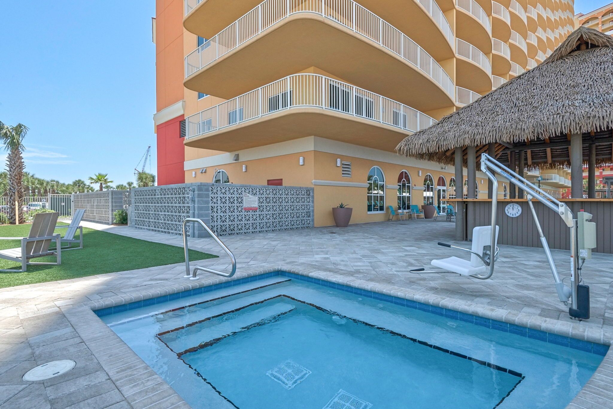 Una piscina con techo de paja, un edificio de varios pisos con balcones y un área cercada con sillones de descanso.