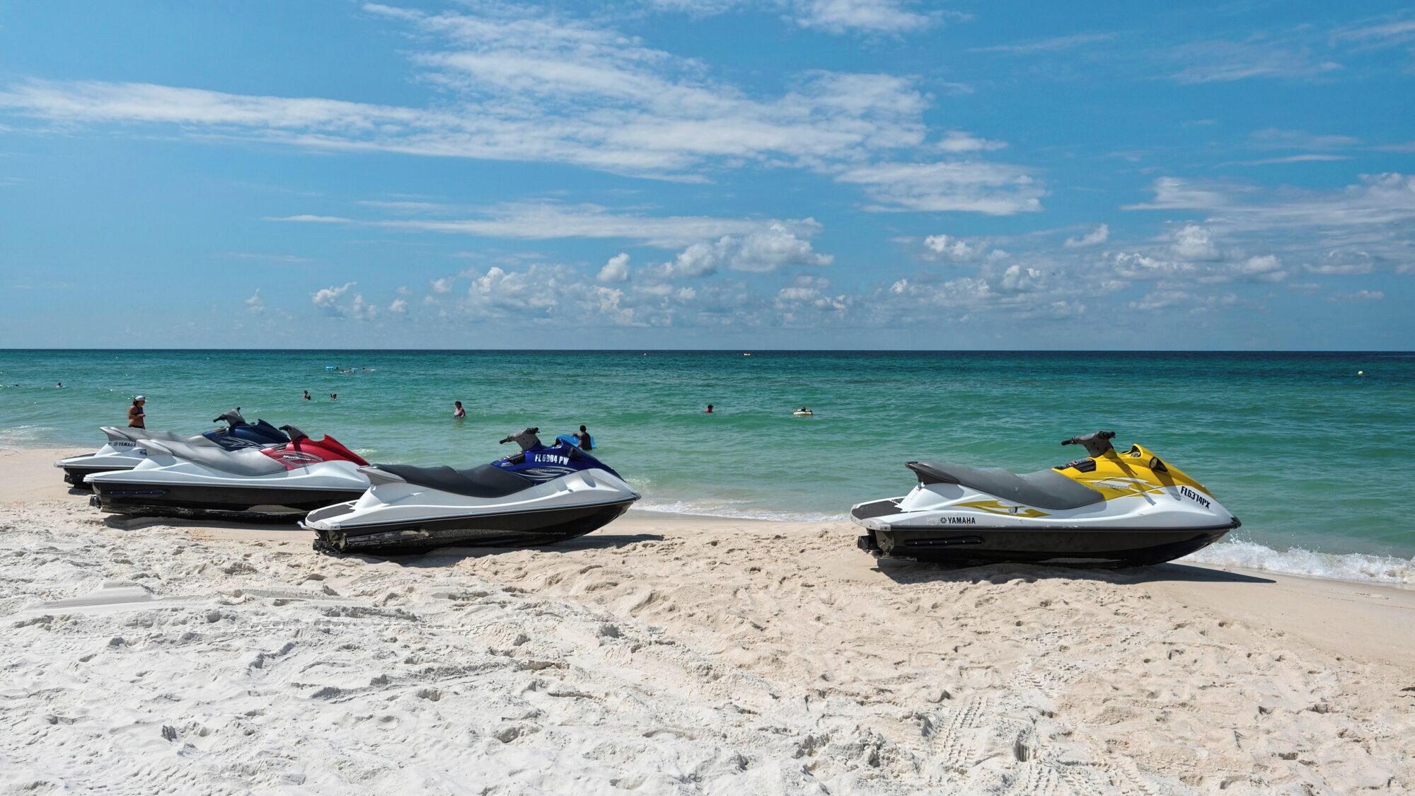 A beach with three jet skis parked on the sand, a clear sky, and the ocean in the background.