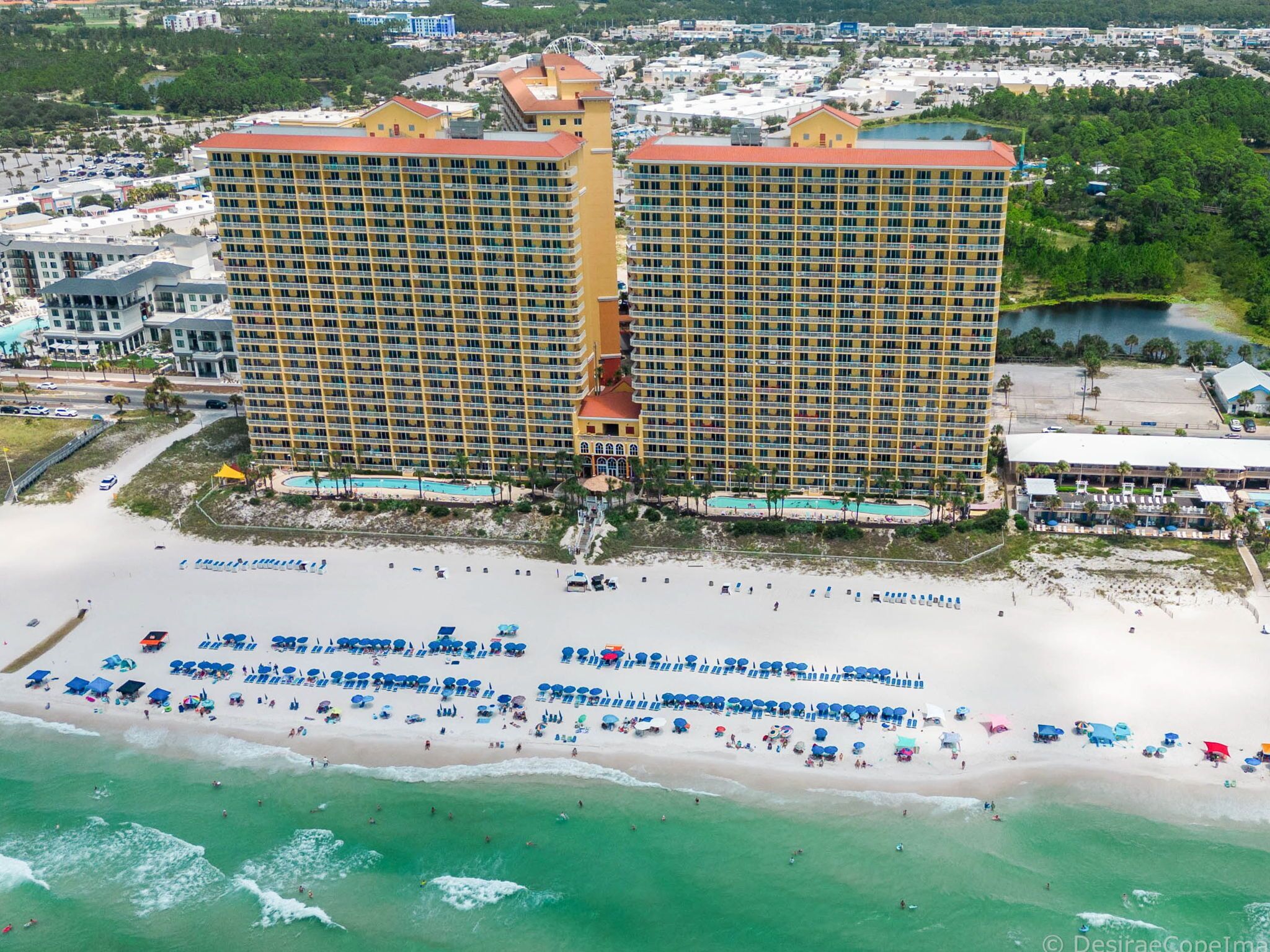 Ein großes Hotel mit mehreren Etagen, ein Strand mit zahlreichen Sonnenschirmen und einem Poolbereich.