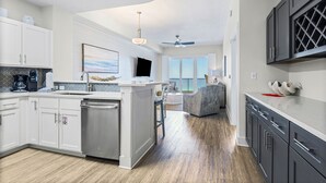 A modern kitchen with a central island, a dining area with a gray sofa, and a view of the outdoors through large windows.