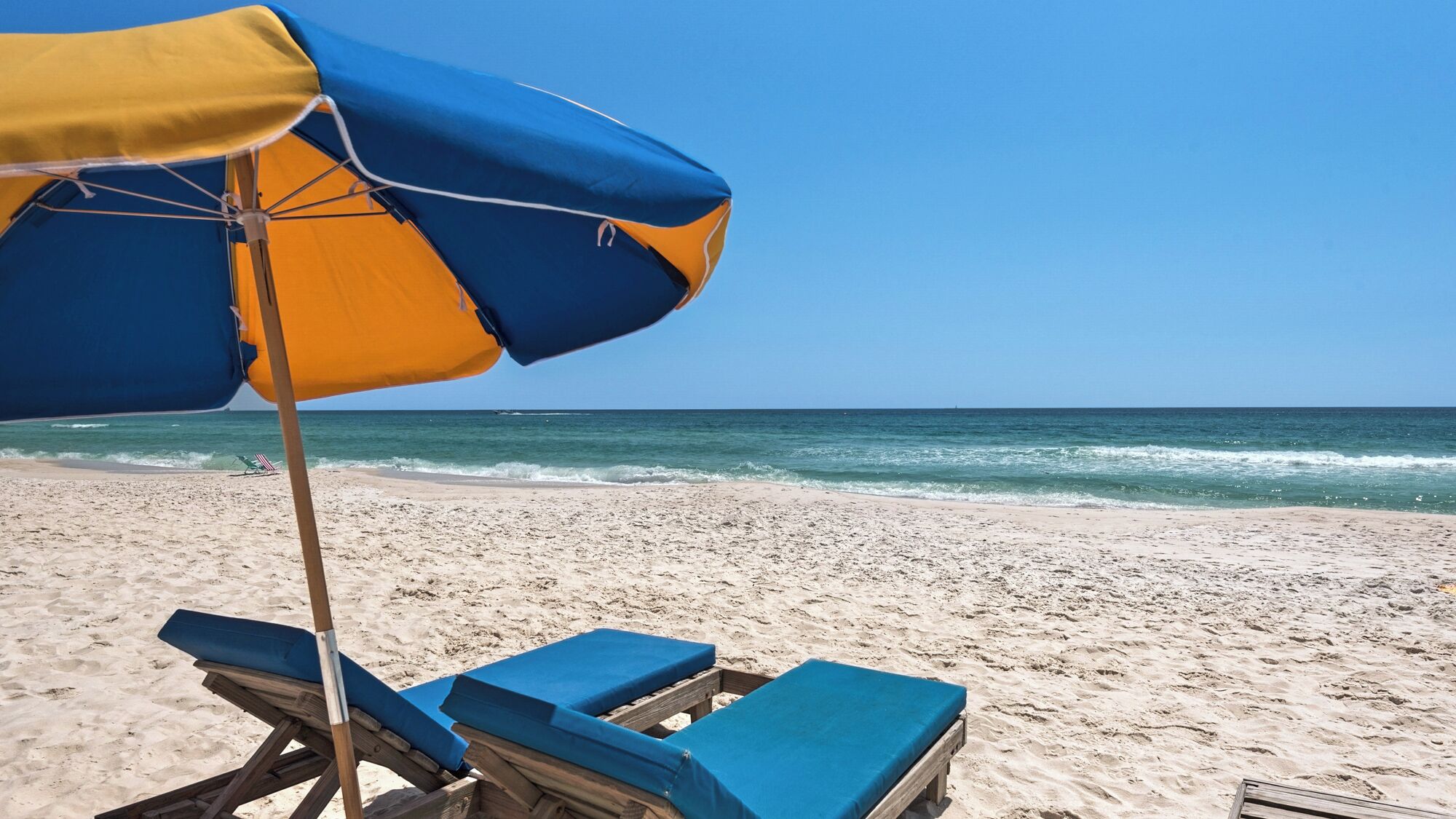 A beach with a blue and yellow umbrella, wooden lounge chairs, and a sandy shore.