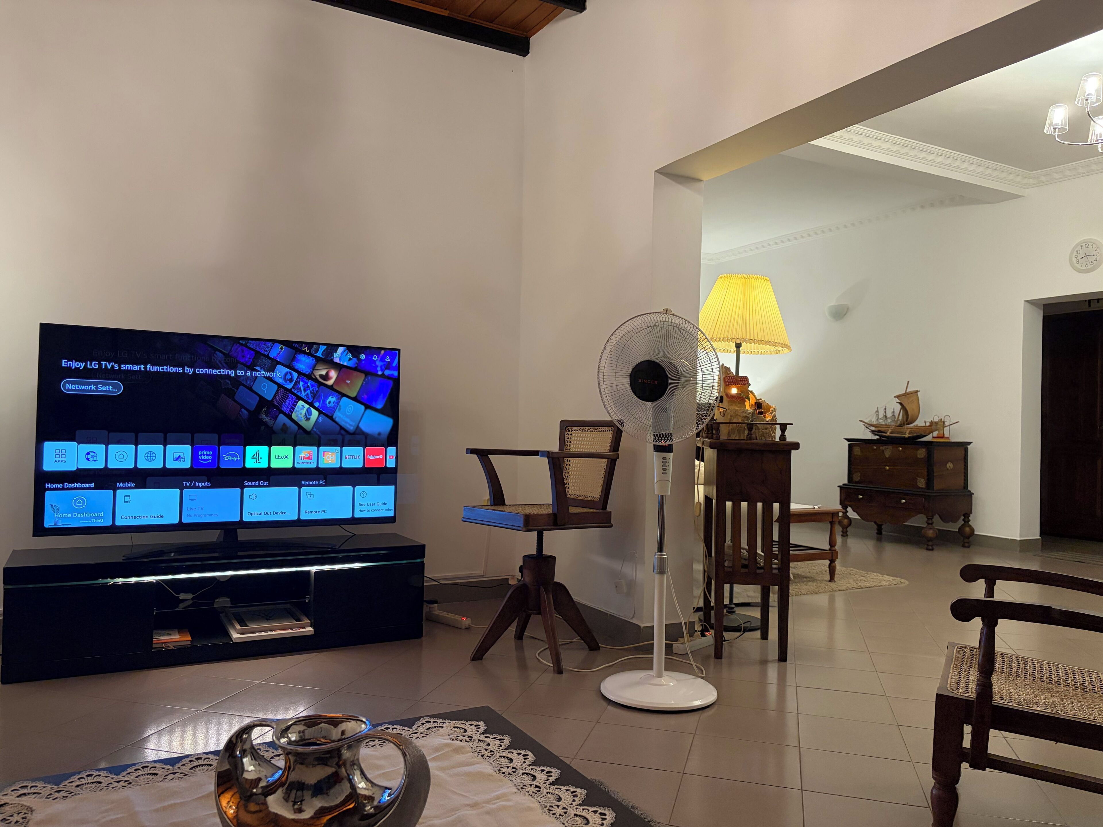 A living room with a TV, a standing fan, a lamp, and a decorative ship model on a dresser.