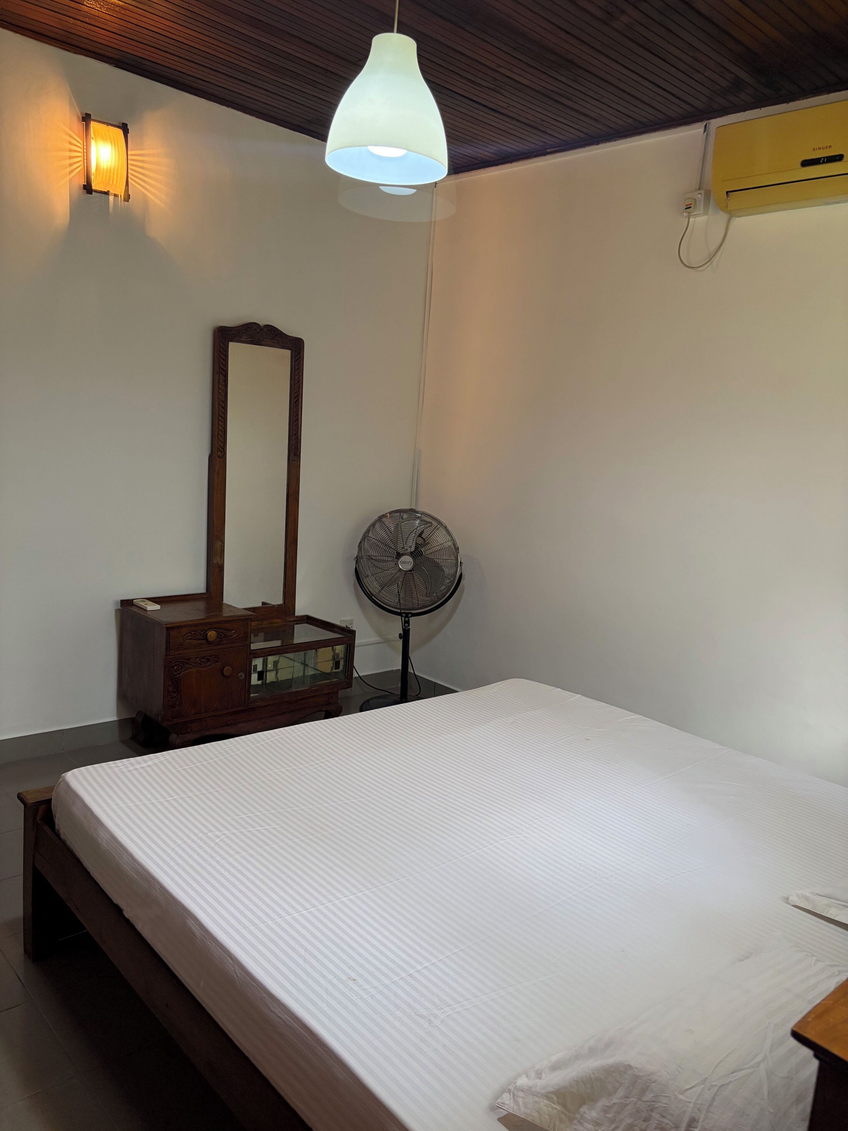 A bedroom with a bed, a wooden dresser, a mirror, a wall-mounted fan, and a ceiling light fixture.