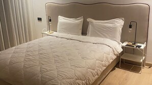 Een net opgemaakt bed met een gewatteerde spreidek, twee nachtkastjes met lampen, en een raam met jaloezieën.