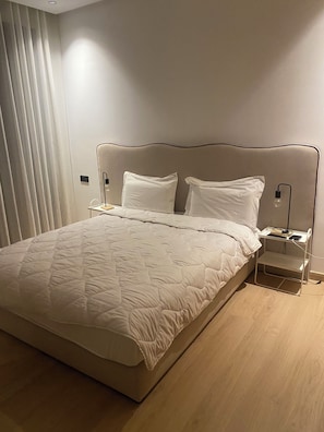 Ein ordentlich bezogenes Bett mit einer gesteppten Decke, zwei Nachttische mit Lampen und ein Fenster mit Jalousien.