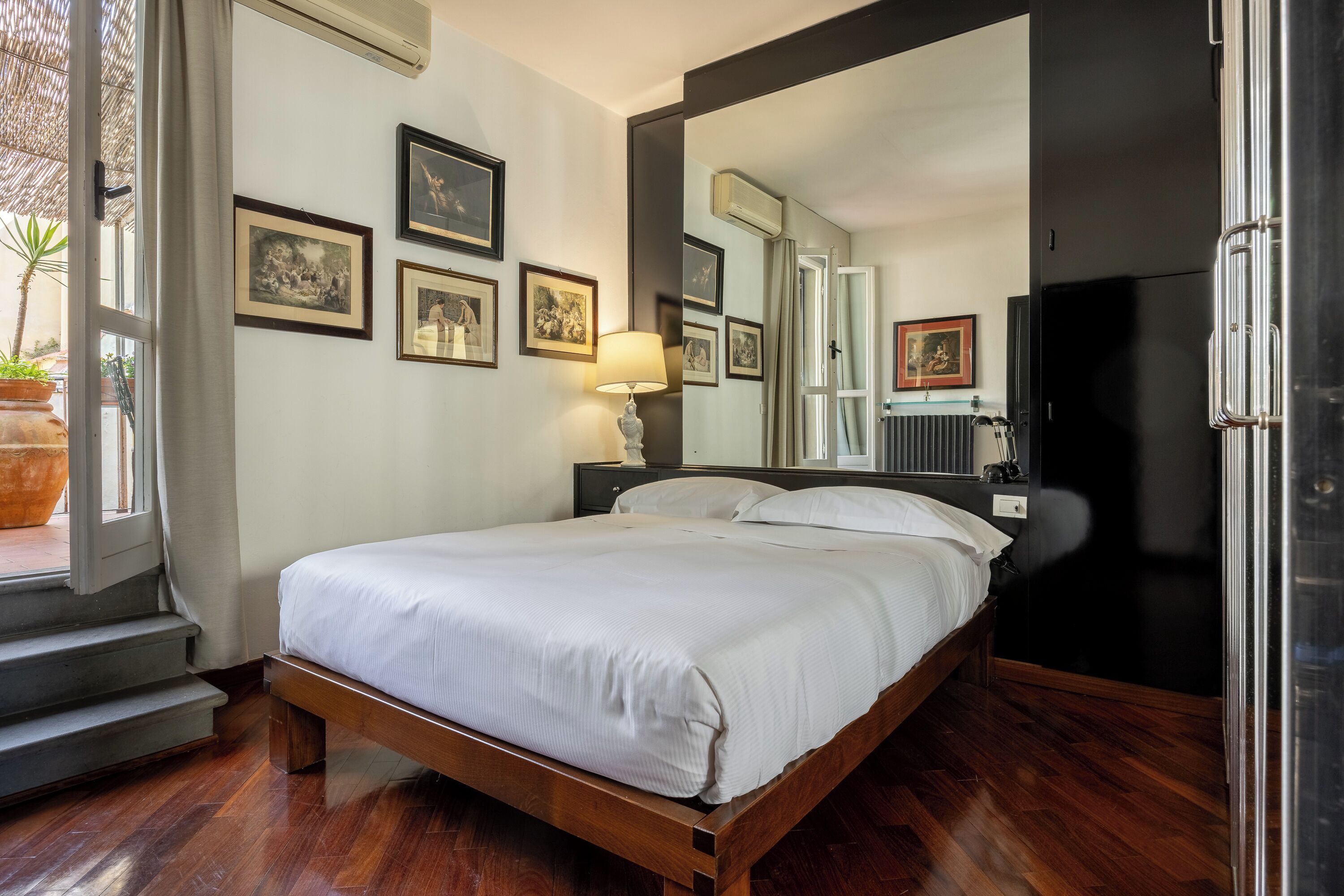 Une chambre à coucher avec un lit, un grand miroir, une fenêtre avec des rideaux et des tableaux encadrés accrochés au mur.