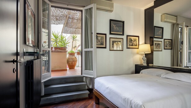 Une chambre à coucher comprenant un lit, une table de chevet, une lampe, un grand miroir et une porte donnant sur un balcon agrémenté de plantes en pot.