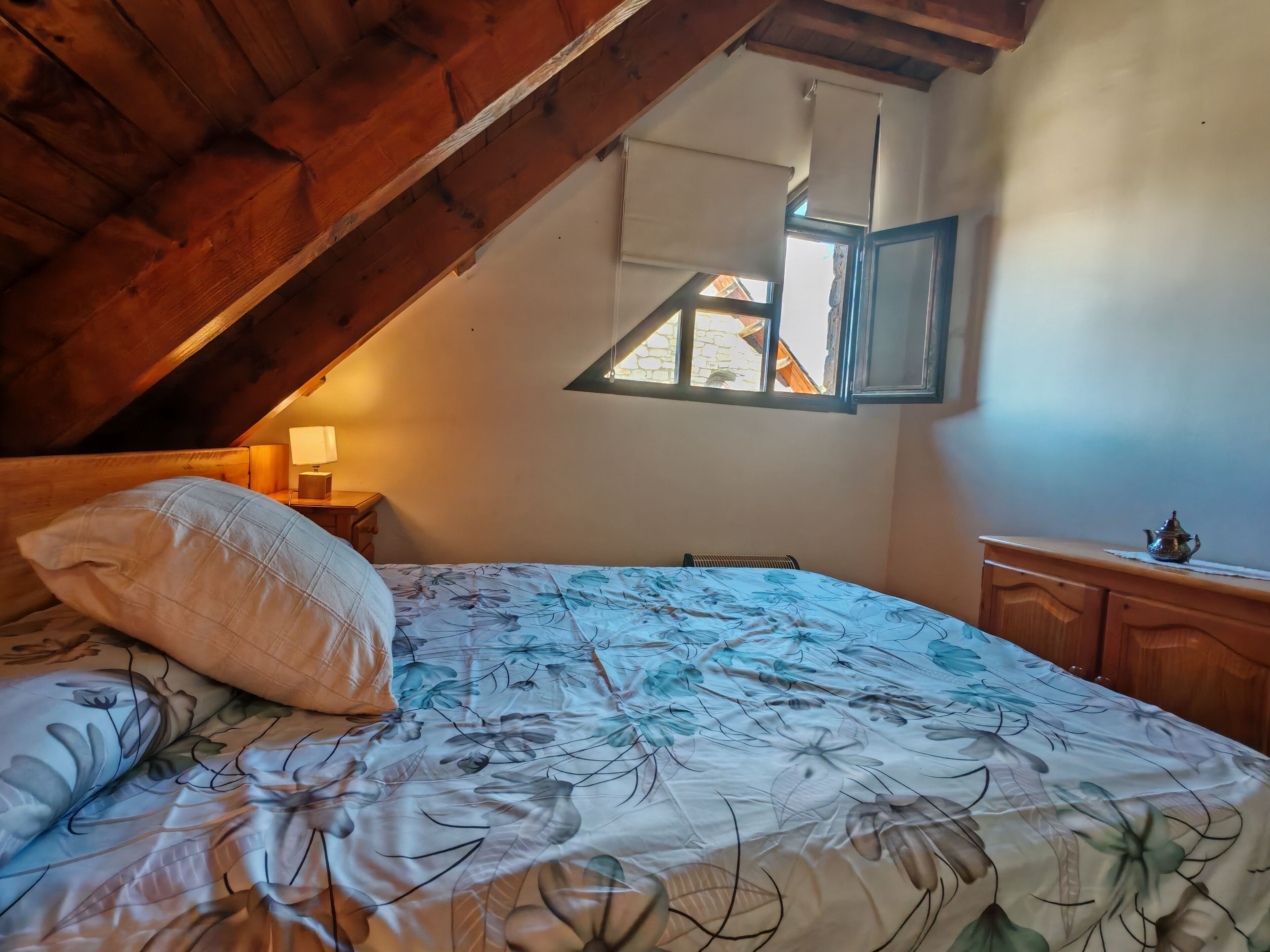 Une chambre avec un plafond en pente, un lit recouvert d’une literie à motifs floraux, une table de chevet avec une lampe et une petite table sur laquelle se trouve une théière.