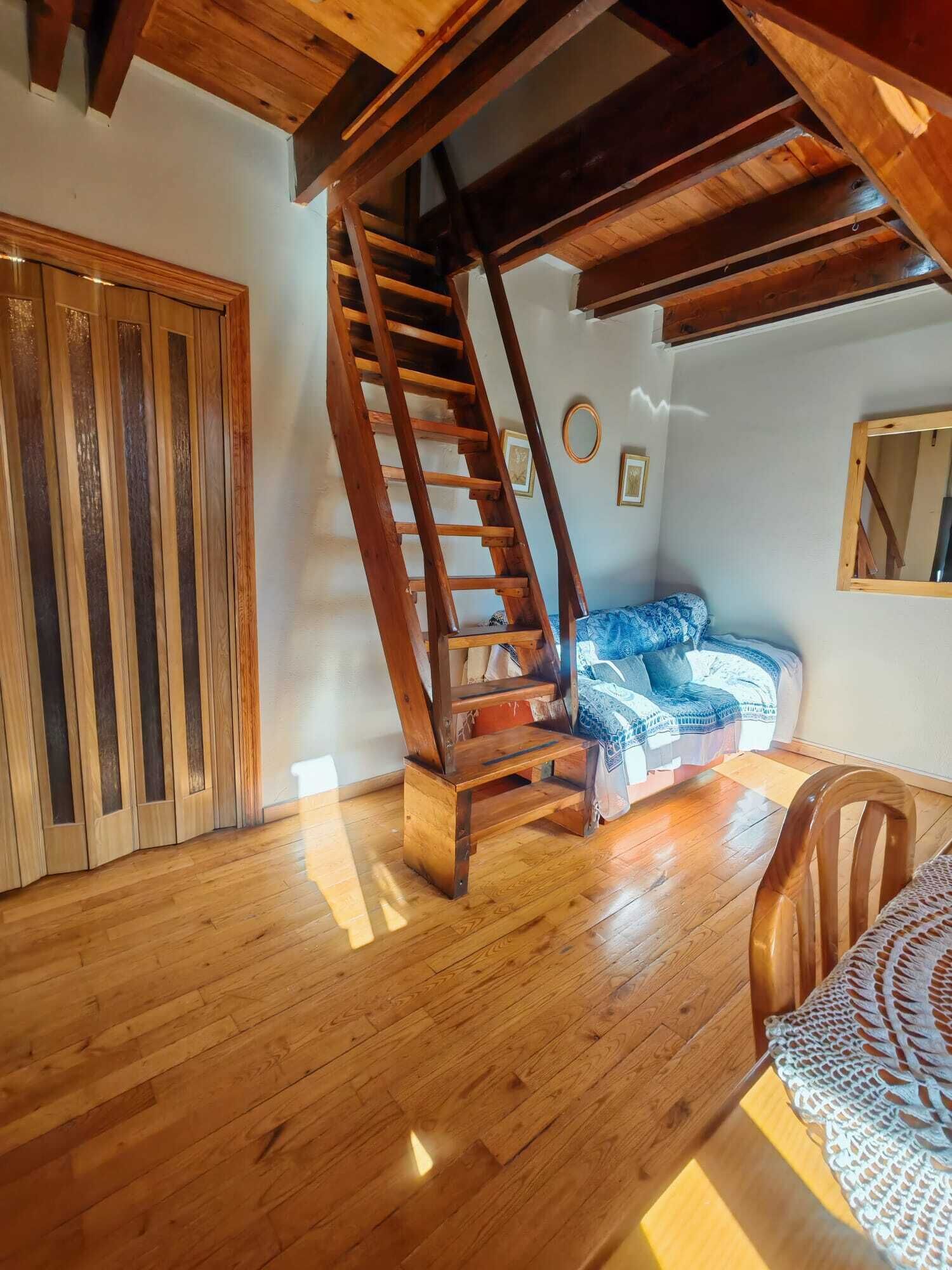 A wooden staircase leading to an upper level, a wooden door, a bed with a patterned blanket, and a wooden chair.