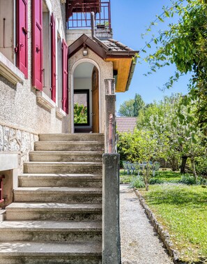 Un escalier en pierre menant à une porte en bois avec des volets rouges, un jardin avec des chaises et un ciel bleu azur.