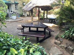A picnic table in a garden with a thatched-roof structure in the background.