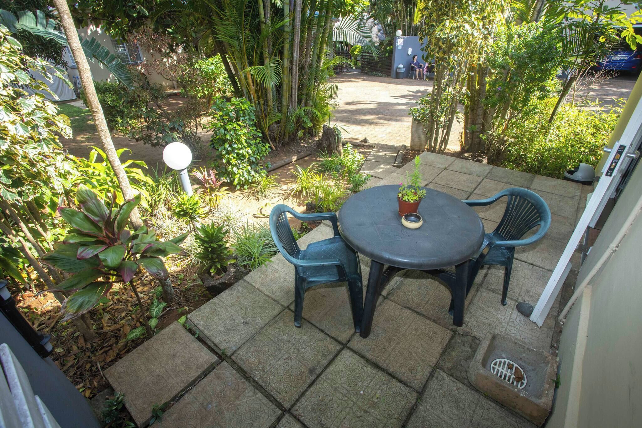A patio with a round table and two chairs, surrounded by various plants and trees.