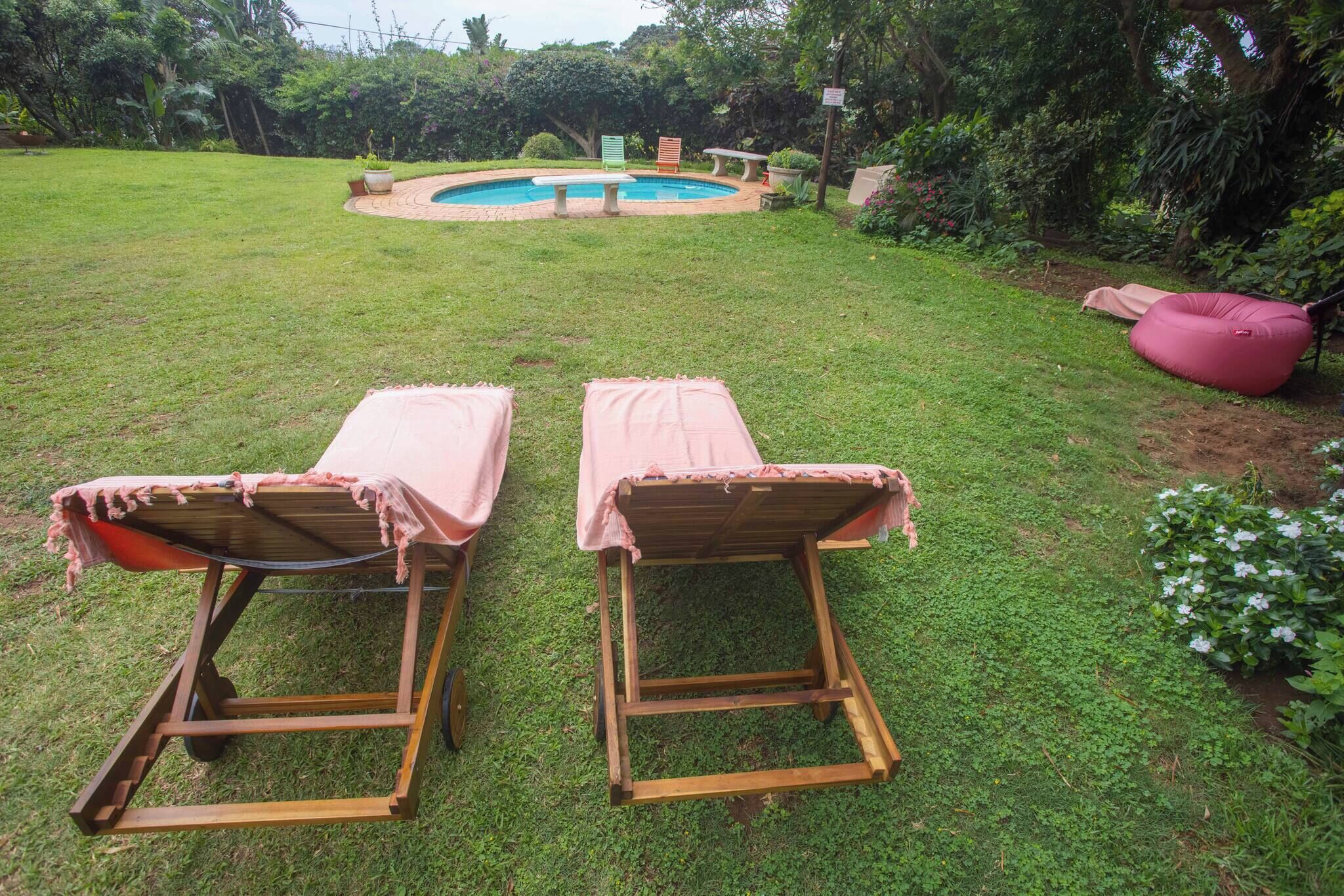 Zwei Holzliegestühle mit rosa Bezügen, ein kleiner Pool und ein Garten mit verschiedenen Pflanzen und einem rosa Sitzsack.