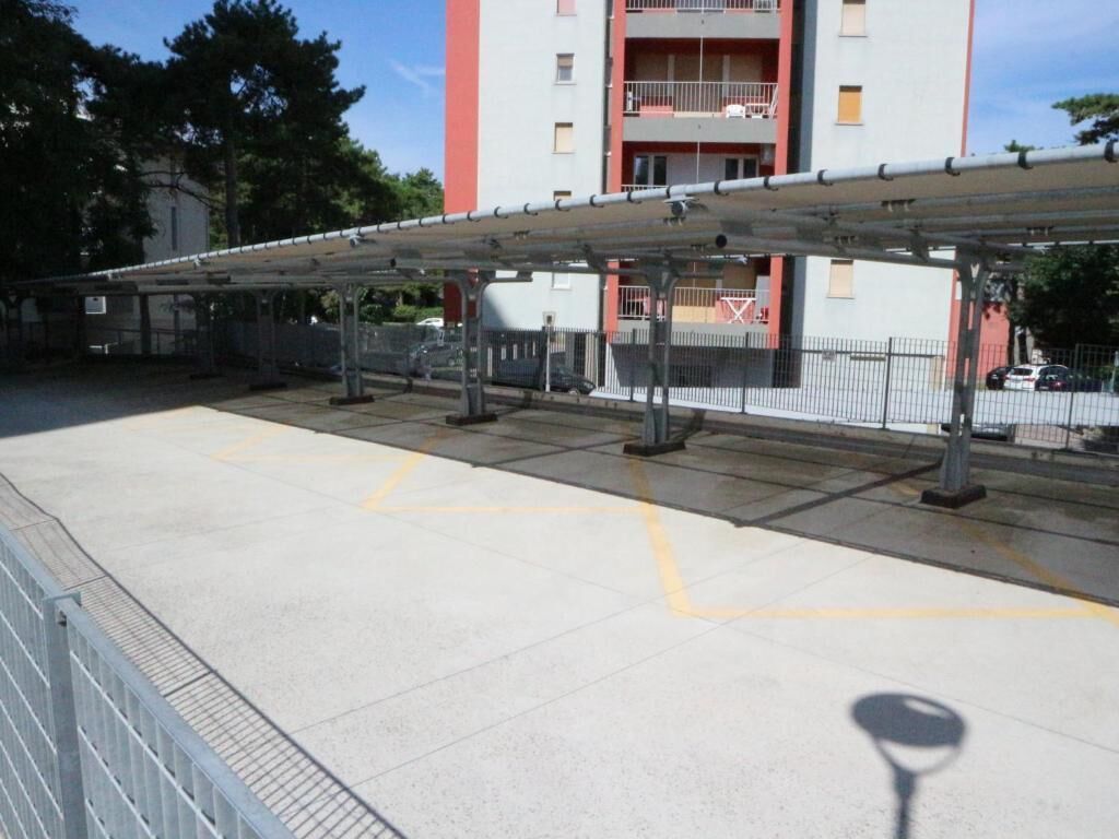A covered pedestrian walkway with a metal railing, a concrete floor with yellow markings, and a multi-story building in the background.