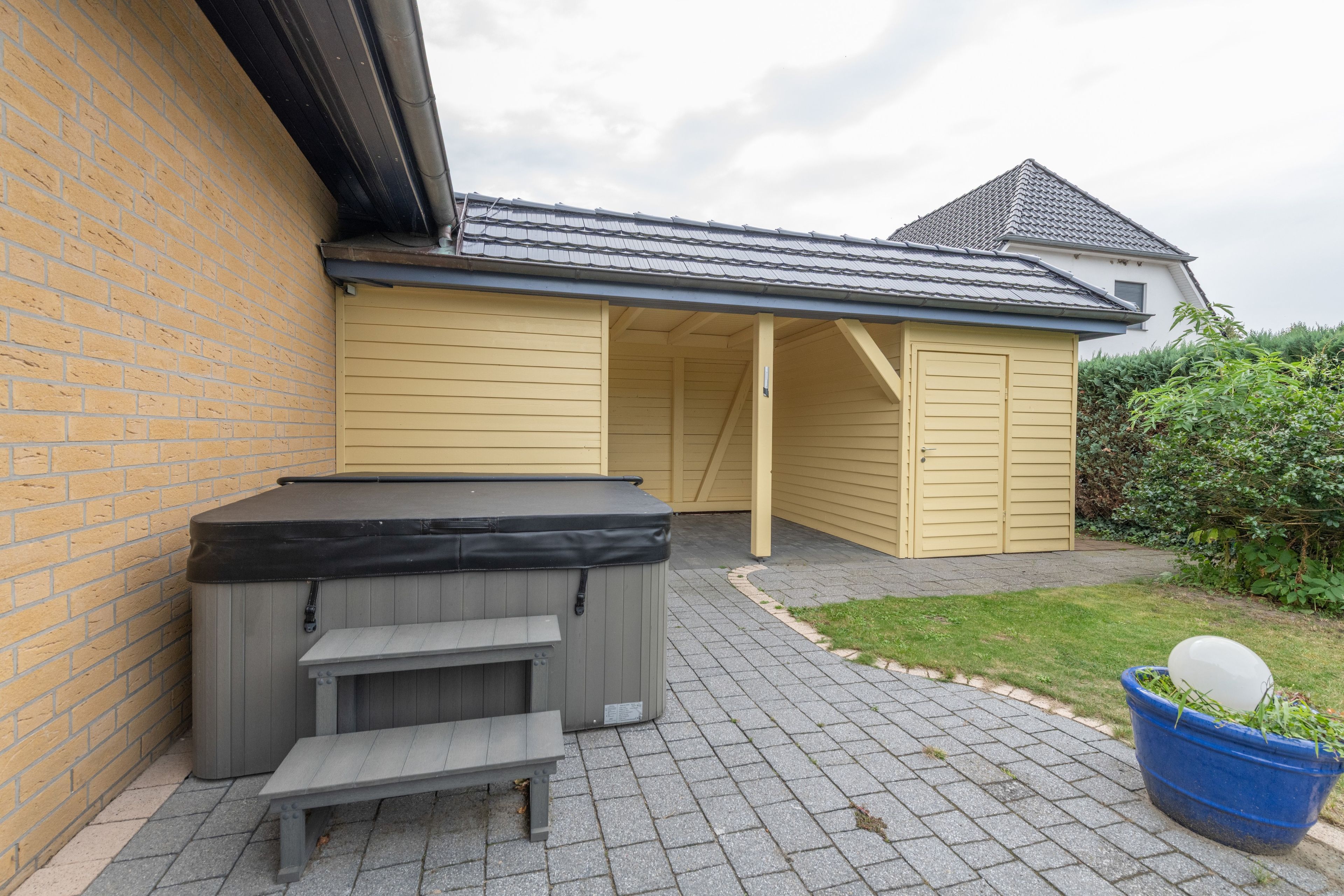 Ein Whirlpool mit Sitzbank, eine überdachte Terrasse und ein blauer Pflanztopf mit einem weißen, kugelförmigen Objekt.