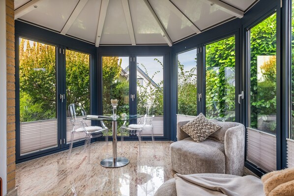 A conservatory with a glass table and chairs, a cushioned armchair, and a view of greenery outside.