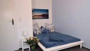A bedroom with a bed, a nightstand, a lamp, a wall-mounted picture, and a vase with flowers. - Bungalow by Lake and Forest for 6 Guests (80 m²) in Untergöhren (Göhren-Lebbin)
