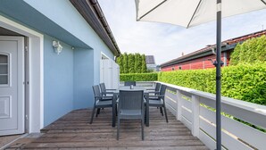Um deck de madeira com mesa e cadeiras, um guarda-sol branco e uma sebe ao fundo.