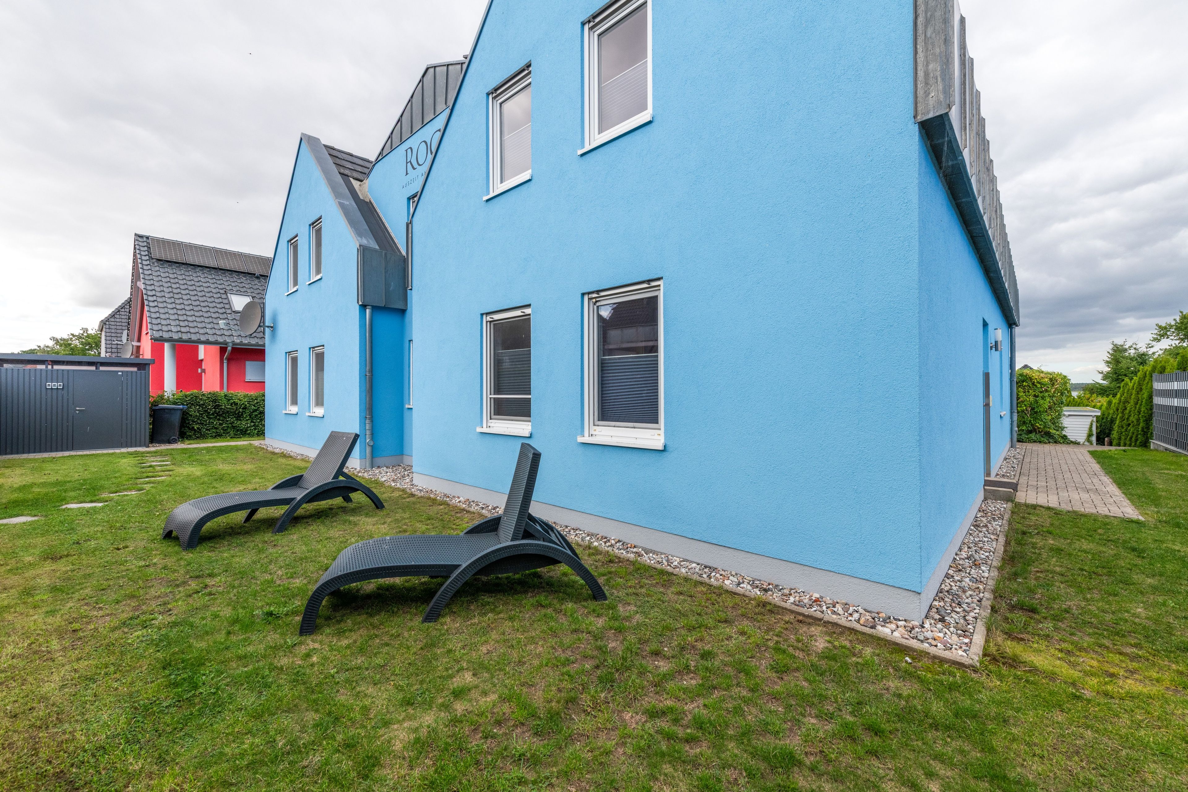 Ein modernes blaues Haus mit geneigtem Dach, ein rotes Gebäude im Hintergrund und zwei schwarze Liegestühle im Vorgarten.