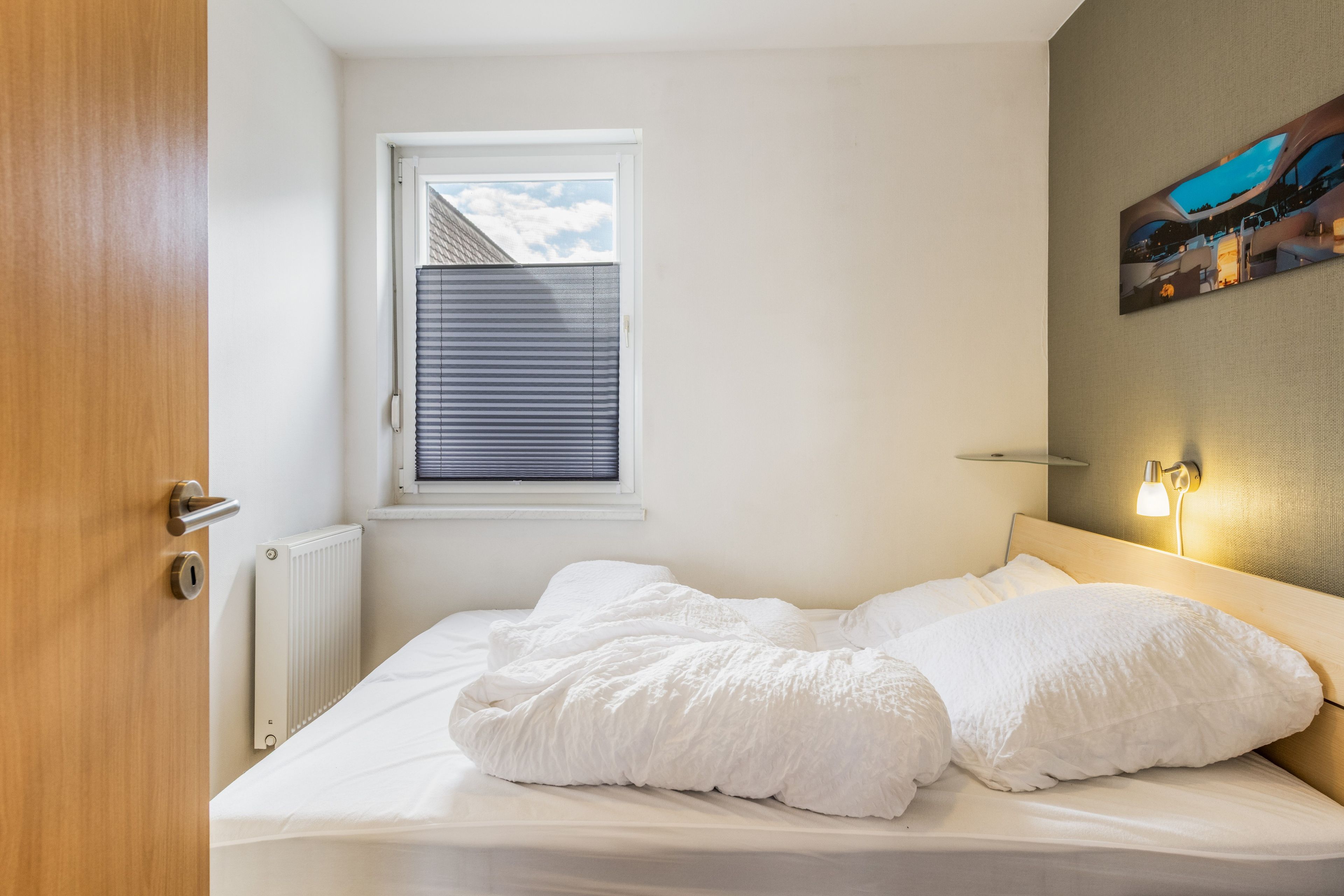 Ein Schlafzimmer mit einem Bett, einem Fenster mit Jalousien, einem Heizkörper und einer Wandleuchte.