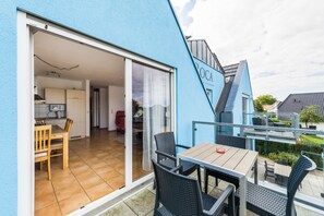 En modern bostadsbyggnad med en blå fasad, en skjutdörr i glas som leder till en matplats med ett träbord och stolar, samt en liten uteplats med bord och stolar.
