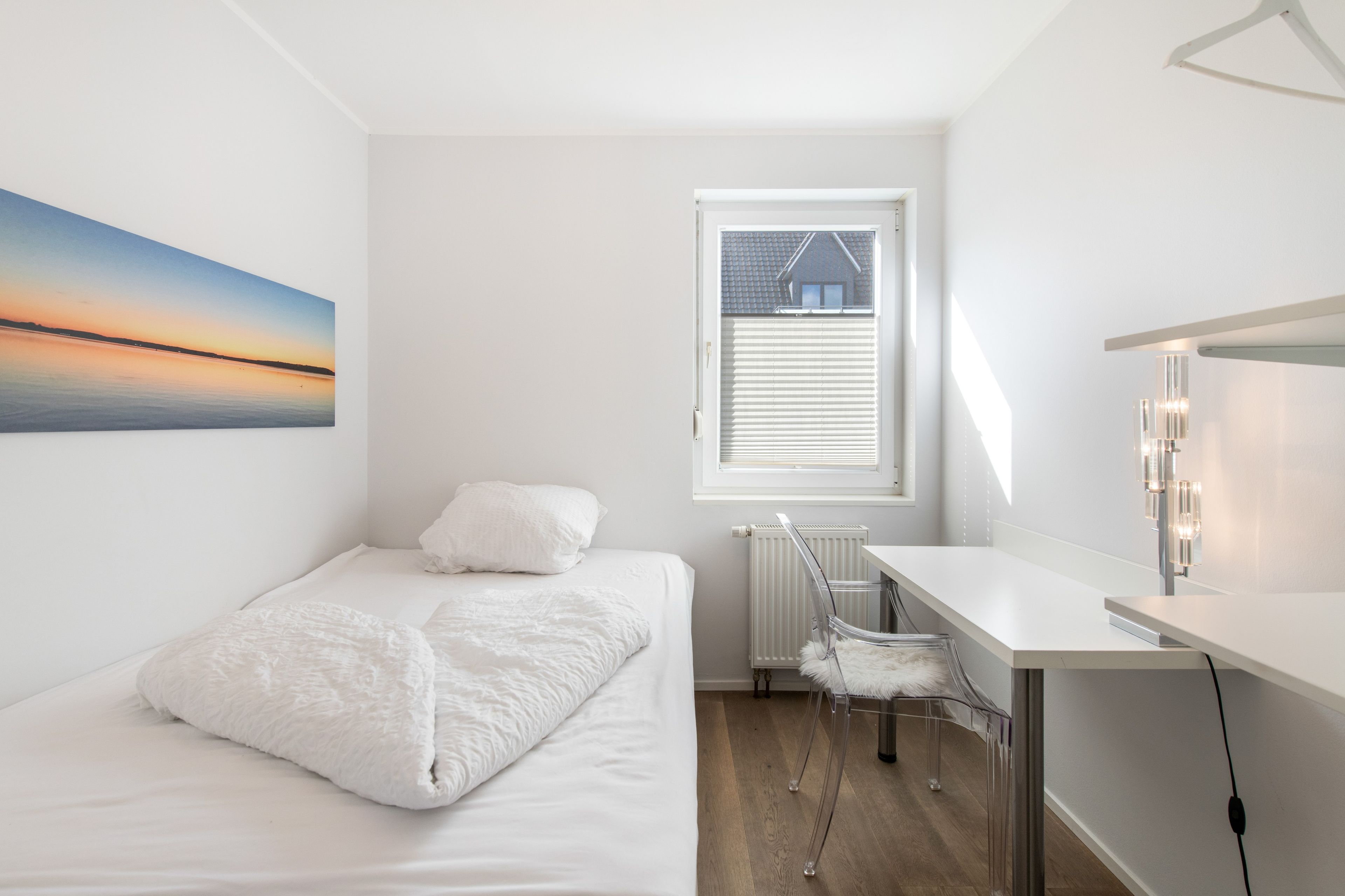 Ein Schlafzimmer mit Bett, Schreibtisch, Stuhl, Heizkörper und einem Fenster mit Jalousien.