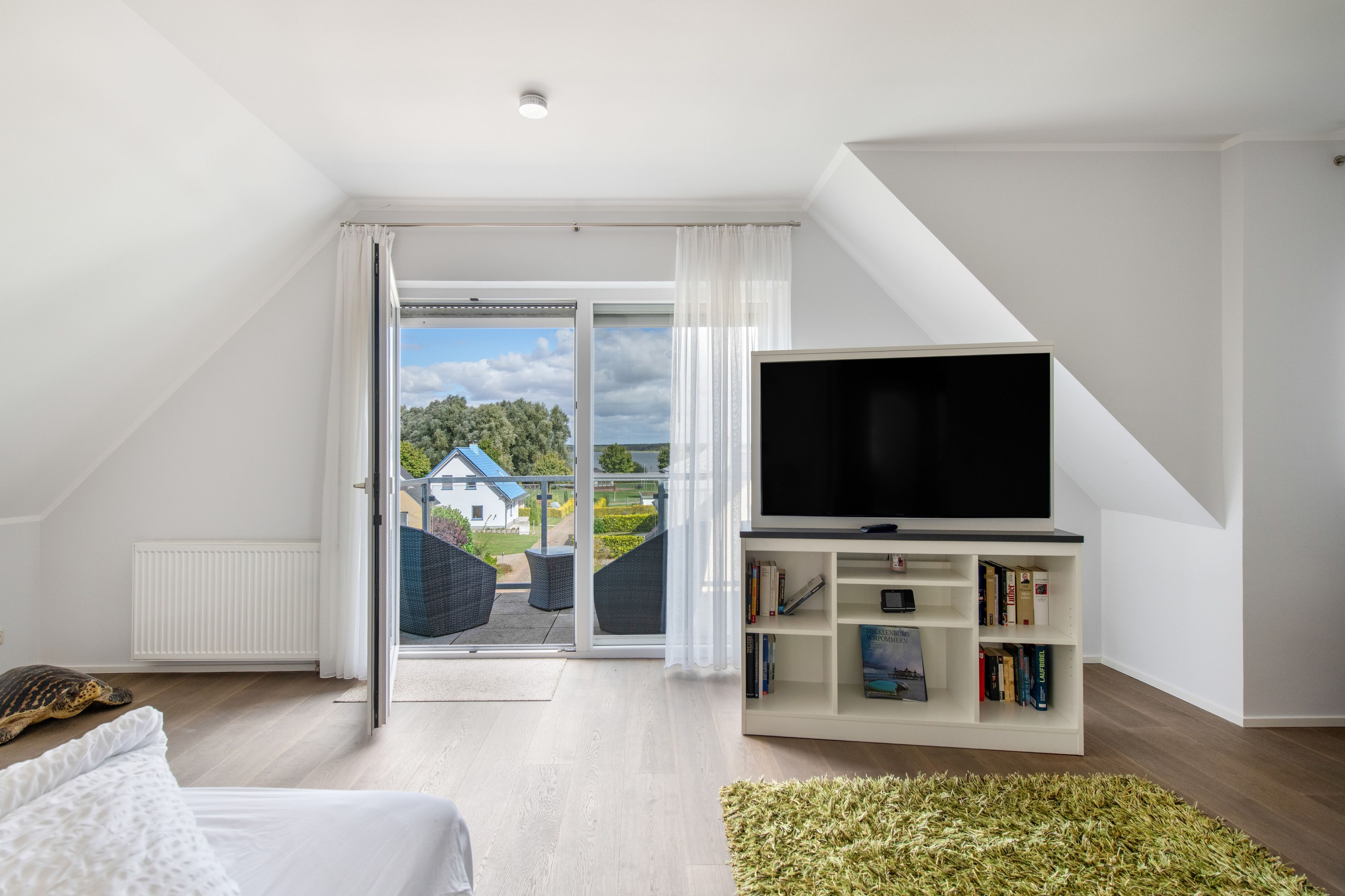 A modern living room with a large TV, a bookshelf, and a view of a house through the sliding glass door.