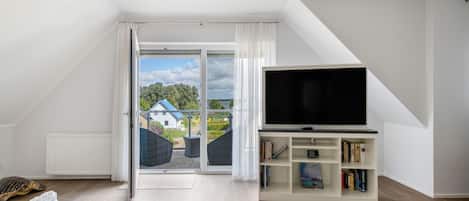 A modern living room with a large TV, a bookshelf, and a view of a house through the sliding glass door.