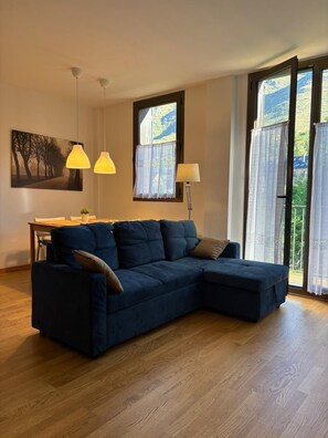 Una sala de estar con un sofá seccional azul oscuro, una mesa de comedor con sillas, lámparas colgantes, un cuadro y vistas a través de un amplio ventanal.