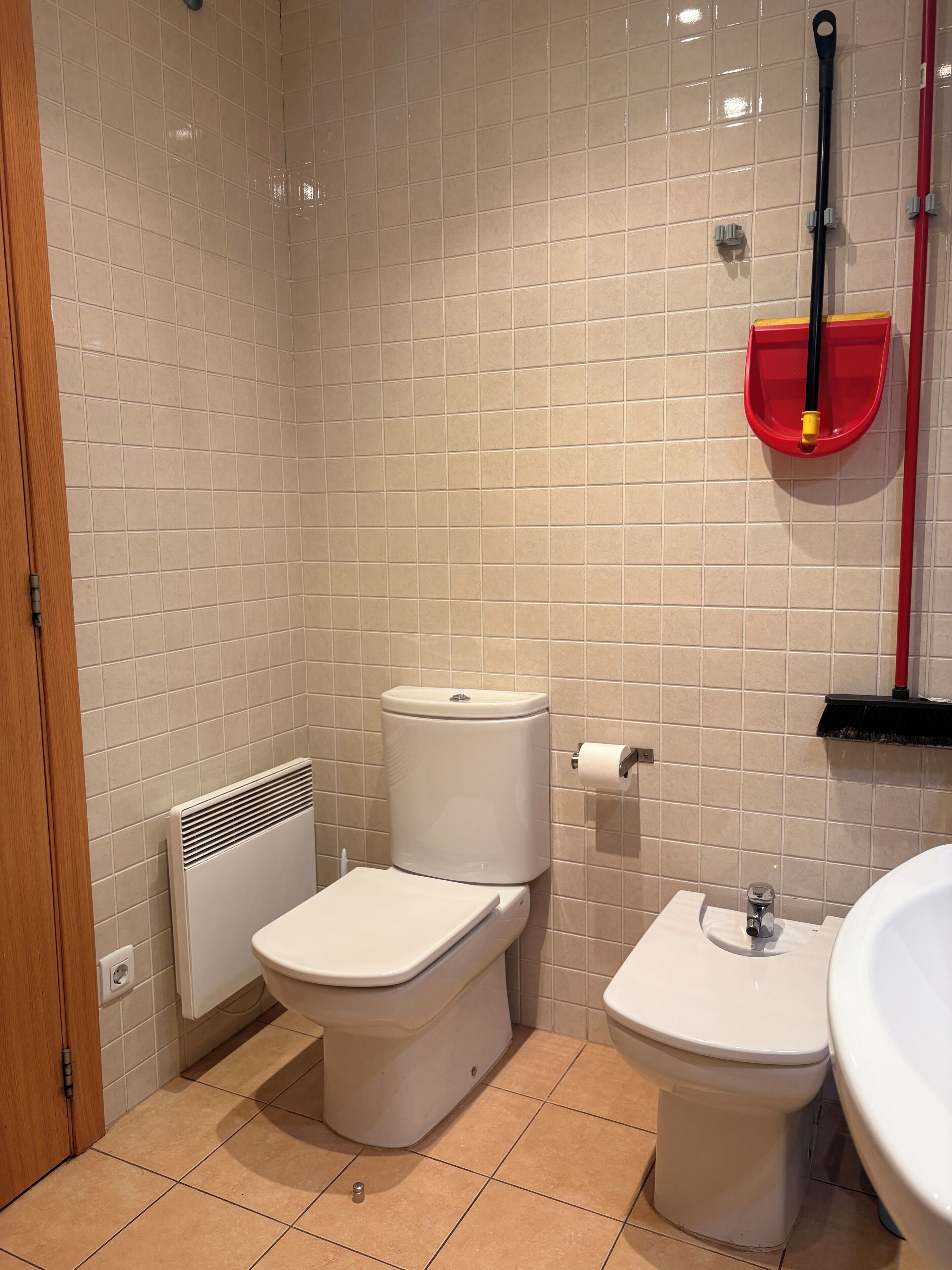 Une salle de bain avec deux toilettes, un radiateur, un balai rouge et un lavabo blanc.