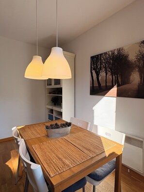 Un'area pranzo con un tavolo di legno, sedie bianche, sottopiatti intrecciati, una ciotola di metallo piena di pigne, lampade a sospensione, un armadietto bianco e un quadro appeso al muro.