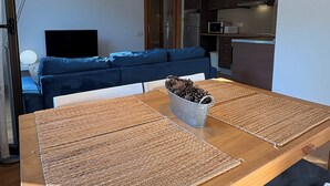 Ett modernt vardagsrum med ett matbord i trä, två vävda bordstabletter, en metallhink med kottar, en blå soffa och en köksdel med skåp och vitvaror.