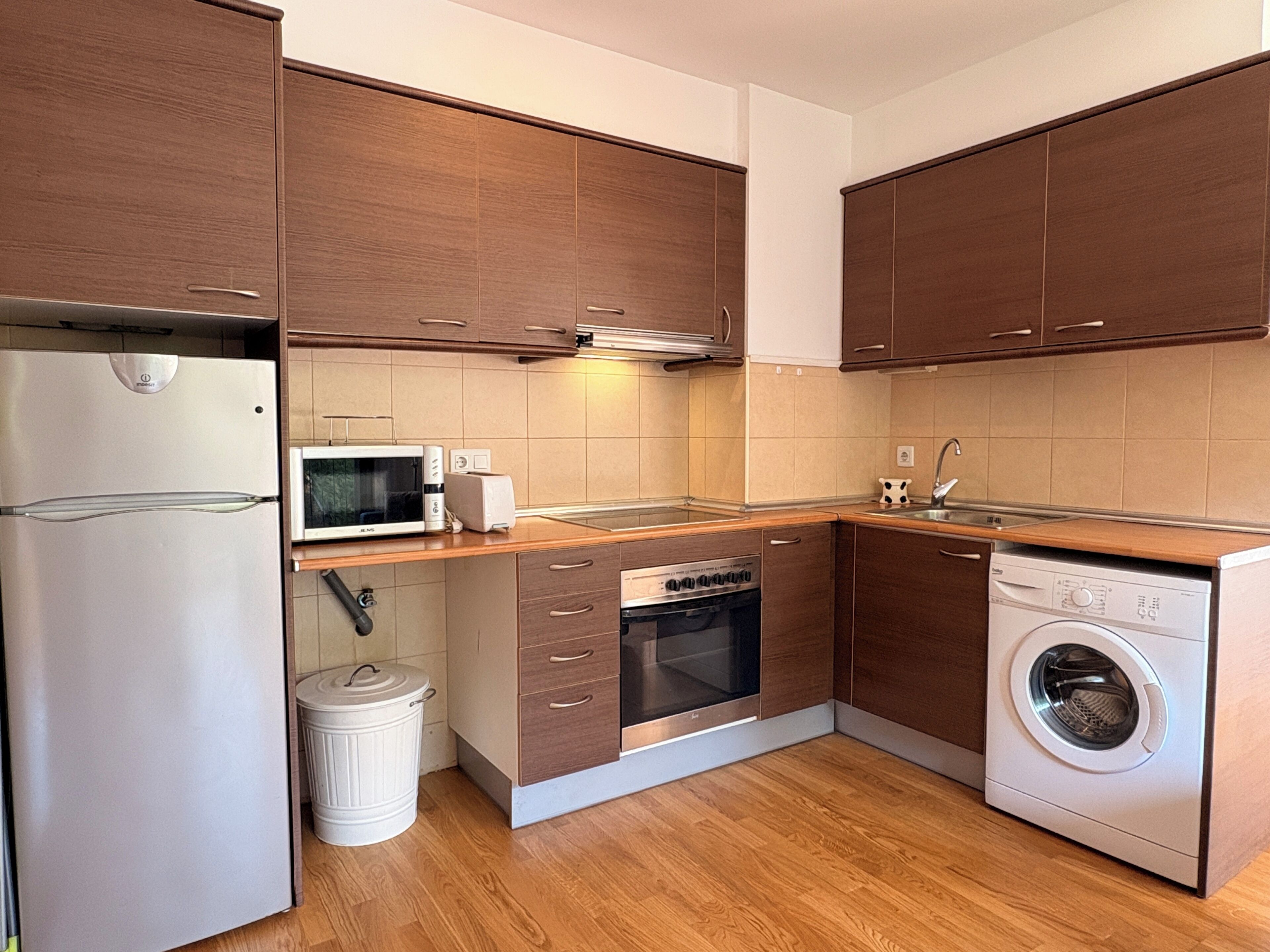 A compact kitchen with wooden cabinets, a stainless steel refrigerator, a microwave, a dishwasher, and a washing machine.