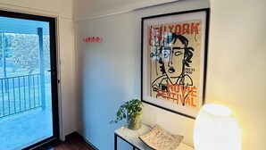 A room with a wooden floor, a framed poster on the wall, a small table with a plant, and a lit lamp.